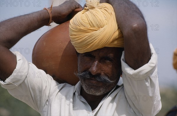 INDIA, Rajasthan, Thar Desert, Bhansda Village.  Portrait of desert man wearing yellow turban and carrying water vessel on his shoulder.
