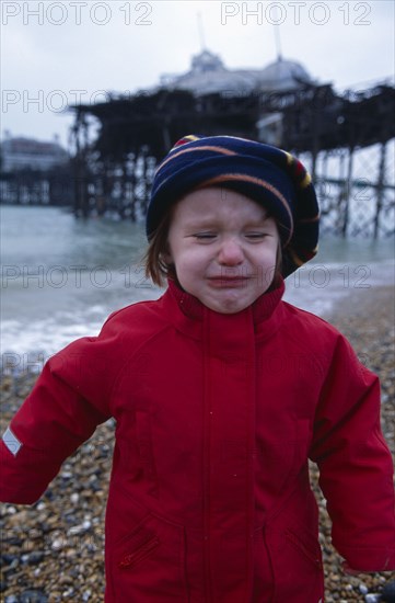 CHILDREN, Playing, Toddler dressed in red coat crying on Brighton Beach with view of the crumbling pier behind.