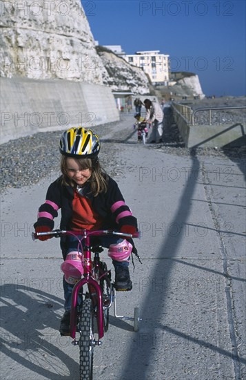 CHILDREN, Playing, Young girl on a bicycle dressed in protective cycling helmet and pads riding on a path next to the beach.