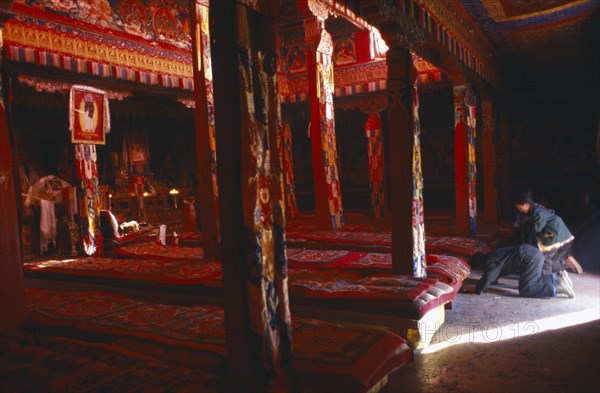 TIBET, Sakya, View through the interior of a temple in red ambient light towards the monks long cushions and Pilgrims prostrating at the entrance.