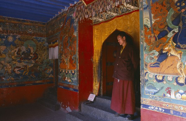 TIBET, Sakya, Monk standing in a doorway with brightly painted religious scenes and deities around him.