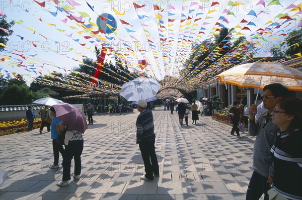 CHINA, Yunnan, Kunming, People walking on a path holding umbrella’s in Daguan Park with colourful flags overhead.
