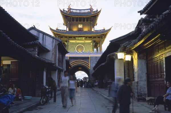 CHINA, Yunnan, Dali., Weishan. View down street at dusk towards an illuminated tower with people strolling up and down.
