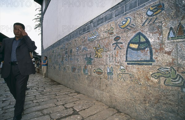 CHINA, Yunnan, Lijiang, Old town Naxi pictogram writing on a wall with a man dressed in a suit walking past on the pavement.