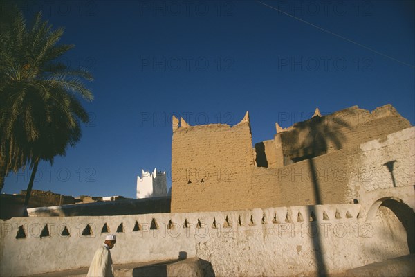 LIBYA, Ghadames Oasis, Man walking past whitewashed wall with mud brick town walls and white mosque minaret behind