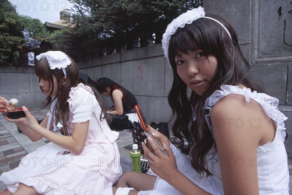 JAPAN, Honshu, Tokyo, Harajuku District. Two teenage girls sitting together with one eating and the other using a mobile phone