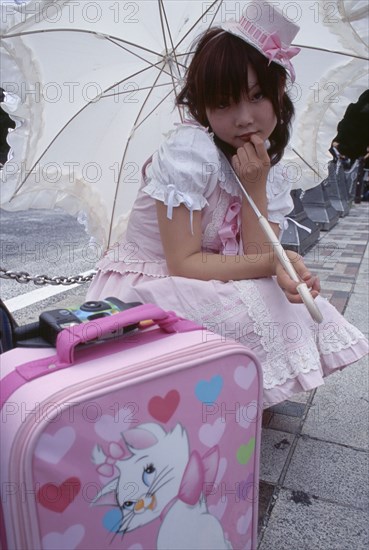 JAPAN, Honshu, Tokyo, Harajuku District. Portrait of young teenage girl wearing a pink and white dress holding a parasol with a pink suitcase in the foreground