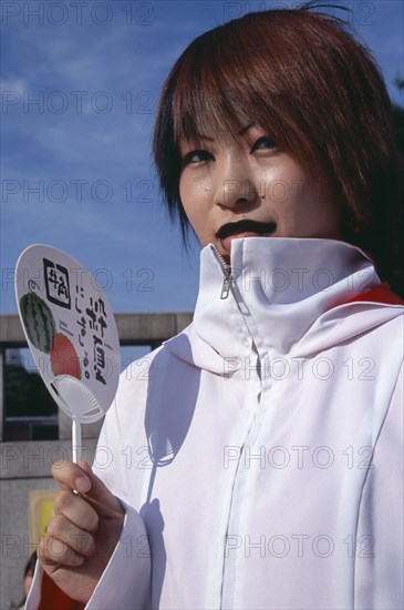 JAPAN, Honshu, Tokyo, Harajuku District. Portrait of a teenage girl wearing a white jacket holding a fan