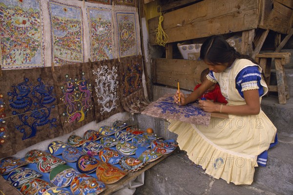 MEXICO, Puebla, Taxco, Young woman painting design on piece of bark with masks and other paintings displayed beside her.