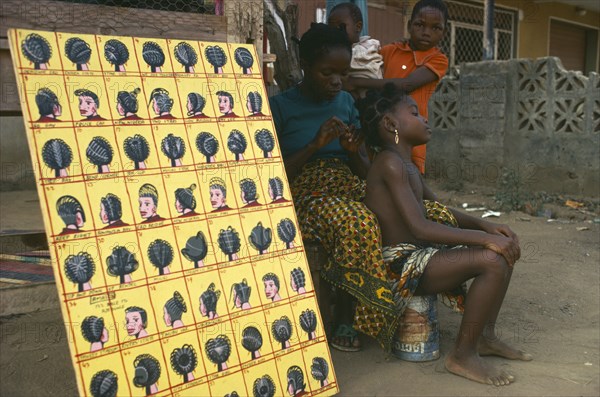 NIGERIA, Lagos, Hairdresser braiding hair of young girl at roadside beside board displaying different hairstyles.