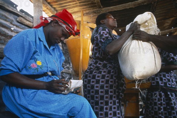 TANZANIA, Food Aid, Women lifting up a sack of food aid next to a woman sat writing on piece of paper.