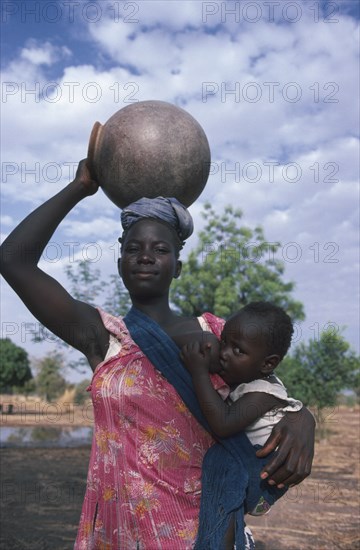 BURKINA FASO, Children, Woman breastfeeding child in sling at her side while carrying water pot on her head.