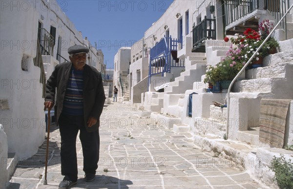 GREECE, Cyclades, Hora, Folegandros.  Street scene with elderly man using stick to walk along pavement lined by white washed houses with steps leading from street to entrance.