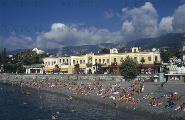 UKRAINE, Crimea, Yalta, People sunbathing on narrow beach or swimming with promenade and waterside buildings behind.