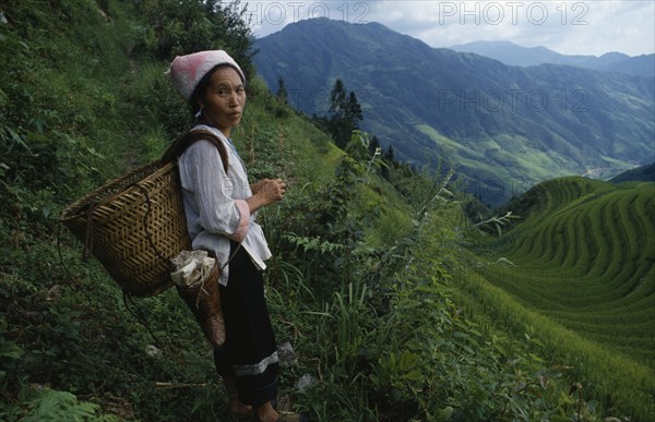 CHINA, Guangxi Province, Longsheng, Woman on steep pathway above rice terraces carrying woven basket on her back.
