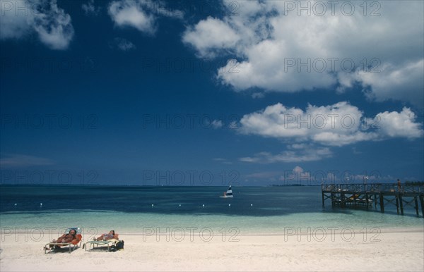 BAHAMAS, Nassau Beach, Two women sunbathing on sandy beach with windsurfer on water beyond.