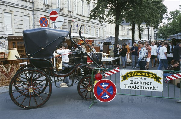 GERMANY, Berlin, Antique carriage at entrance to flea market with people at stalls behind and market and road signs on crash barrier in foreground.