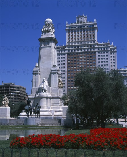 SPAIN, Madrid State, Madrid , Plaza Espana. Stone obelisk with statue of Miguel de Cervantes surrounded by trees and flower beds with tall office building behind.