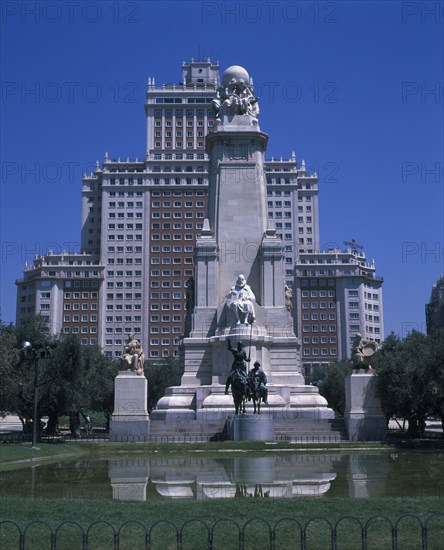 SPAIN, Madrid State, Madrid , Plaza Espana. Stone obelisk with statue of Miguel de Cervantes seen from across pond surrounded by trees with tall office buildings behind.