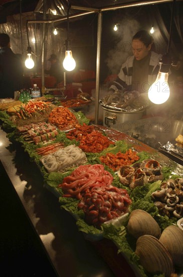 KOREA, Seoul, "Namdaemun Market on a cold December night with a woman cooking behind trays of fresh seafood, pork, chicken and beef with plastic sheeting to keep out cold"