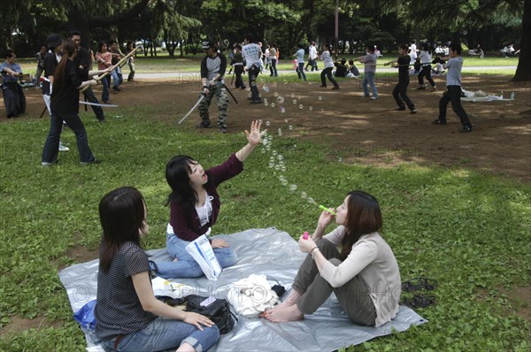 JAPAN, Honshu, Tokyo, "Harajuku. Yoyogi Park. Miki Tomiyoka, aged 23, blows bubbles for two friends while a group practices with swords in the background on a Saturday afternoon"