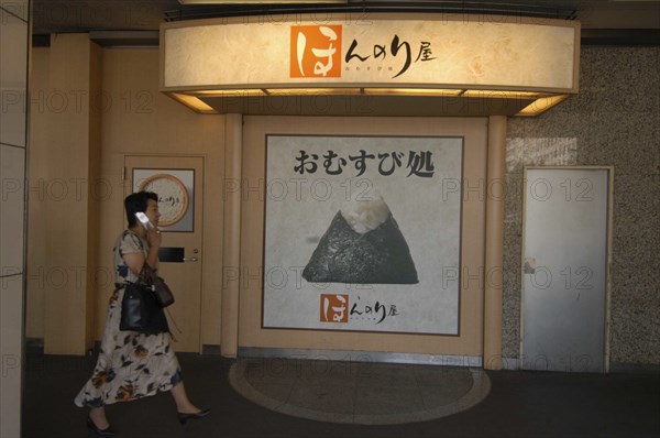 JAPAN, Honshu, Tokyo, Onigiri rice ball shop sign in front of Tokyo Station with passing woman using cell phone