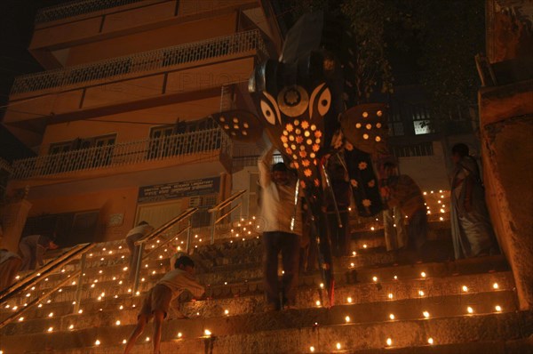 INDIA, Uttar Pradesh, Varanasi, Deep Diwali Festival with men carrying paper statue of Hindu God Ganesh down steps leading to the Ganges River while small boy lights oil lamps