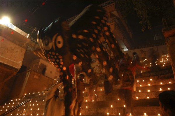 INDIA, Uttar Pradesh, Varanasi, Deep Diwali Festival. Men carry a paper statue of the Hindu God Ganesh down steps covered in oil lamps leading to the Ganges River