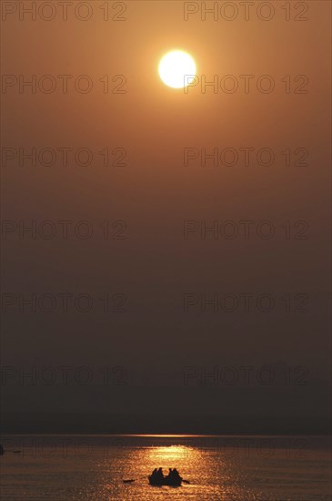 INDIA, Uttar Pradesh, Varanasi, Ganges River at dawn with rowing boat silhouetted against warm sunlight reflected in the water
