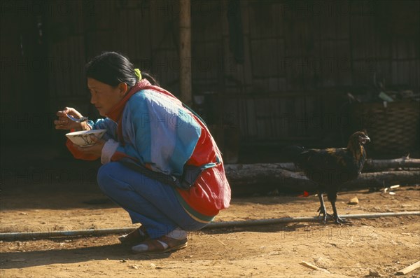 THAILAND, Chiang Rai Province, Doi Lan, Lisu woman eating whilst squatting outside her home with a chicken behind her