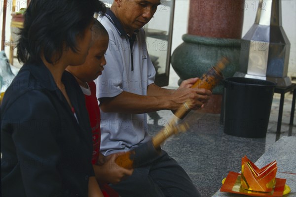 THAILAND, Chiang Mai, Wat Chai Mong Kon, Man with son kneeling before an altar shaking containers of fortune sticks