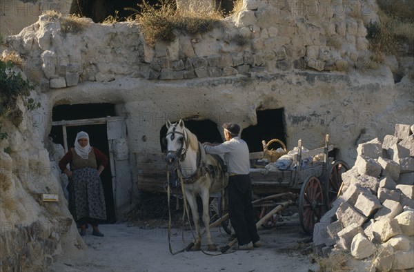 TURKEY, Cappadocia, Uchisar, Couple harnessing pony to wooden cart.  Inhabitants of cave dwellings built into rock behind.