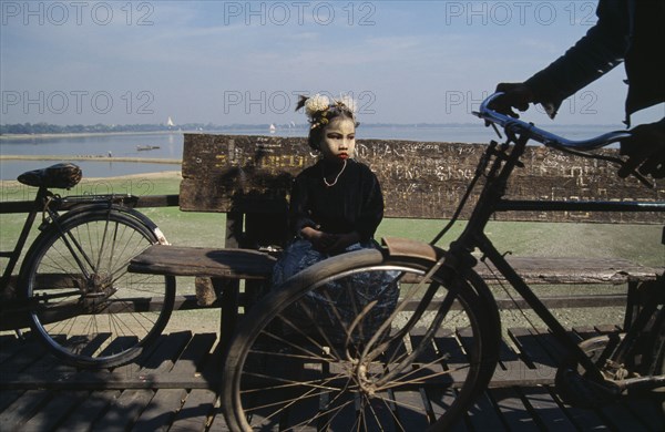 MYANMAR, Lake Taungthaman , Young girl with face decorated with leaf patterns sitting on wooden bench on U-Bein bridge with wheels of passing cyclist in the foreground.