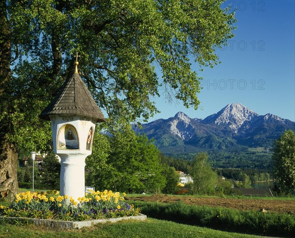 AUSTRIA, Karnten, Egg, Shrine with wooden tiled roof and alcoves painted with religious subjects in rural landscape with mountain backdrop.