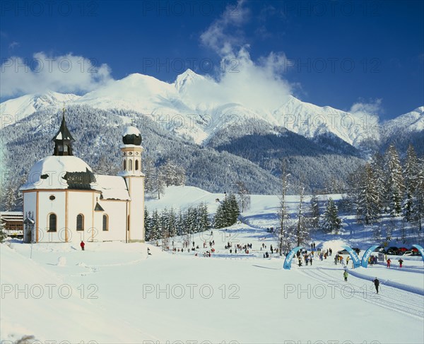 AUSTRIA, Tirol, Seefeld, Church in winter landscape with skiing competition taking place in middle foreground and snow covered mountain backdrop.