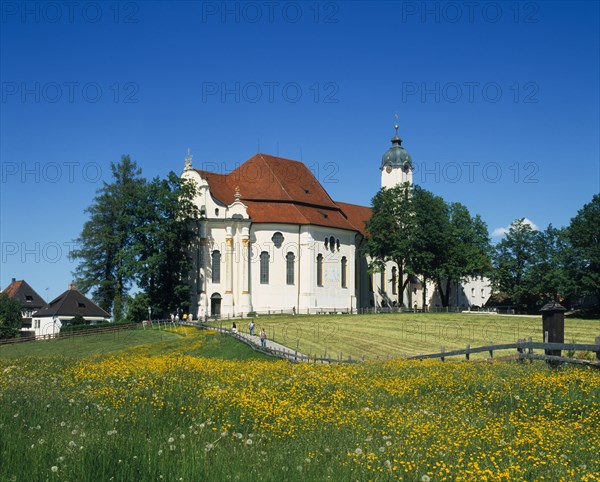 GERMANY, Bayern, General, Visitors on road to white painted church with red tile roof and domed clock tower near Steingaden with field of buttercups in the foreground.