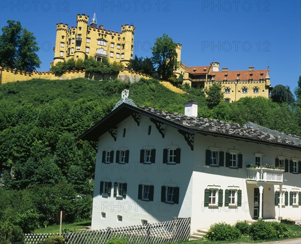 GERMANY, Bayern, Hohenschwangau, Hohenschwangau Castle on hillside overlooking white painted building with window shutters and balcony.
