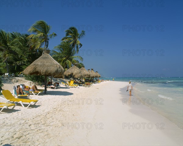 MEXICO, Quintana Roo, Tulum, Stretch of sandy beach with people on sun loungers under a line of thatched sunshades and woman walking along waters edge.