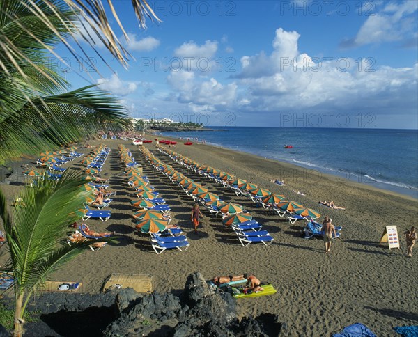 SPAIN, Canary Islands, Lanzarote, Puerto del Carmen.  Sandy beach with lines of blue sun loungers and orange and green parasols for hire.  Few people sunbathing and hotels beyond.