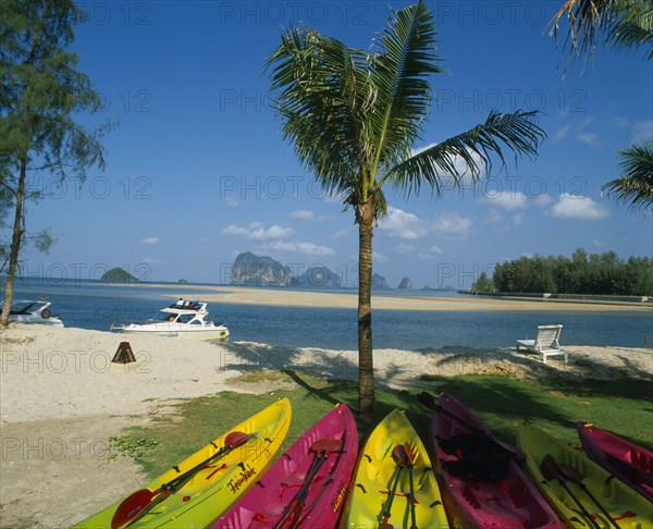 THAILAND, Trang Province, Trang Beach, Brightly coloured canoes under palm tree in foreground with view over white sand beach towards karst formations beyond.
