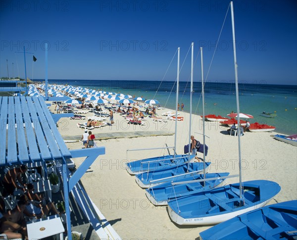TUNISIA, Port el Kantaoui, Busy sandy beach with tourist boats and pedelos for hire and lines of blue and white sun umbrellas.