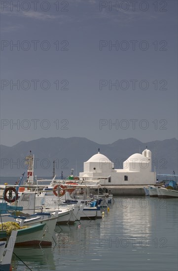 GREECE, Saronic Islands, Aegina, Fishing boats moored in Aegina Town next to a small white church with the mainland in the distance.