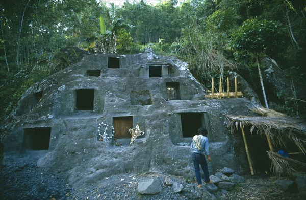 INDONESIA, Toraja, Stone burial ground surrounded by trees with man in the foreground.