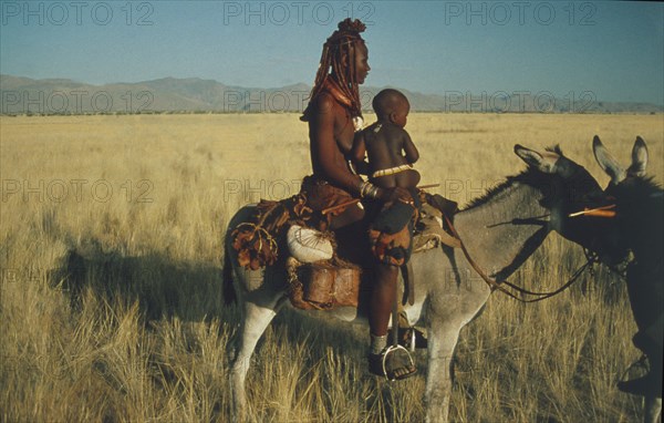NAMIBIA, Kaokolan, Marienfluss, Himba nomad woman and child in traditional leather clothing crossing the Marienfluss on donkey in Kaokolan