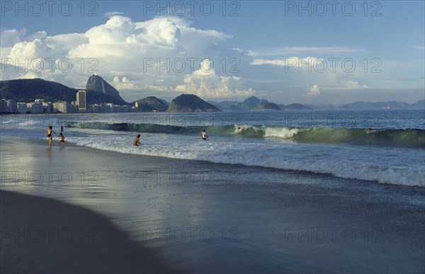 BRAZIL, Rio de Janeiro, Beach with people playing in the surf.  City buildings and Sugar Loaf Mountain beyond.