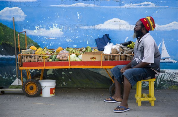 WEST INDIES, St Vincent & The Grenadines, Union Island, Fruit and vegetable stall holder in front of wall painting in Clifton