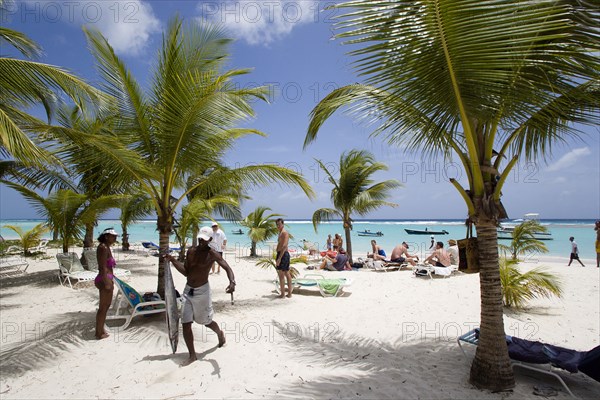 WEST INDIES, Barbados, Christ Church, Fisherman carrying gutted King Fish past tourists and coconut trees on Worthing Beach