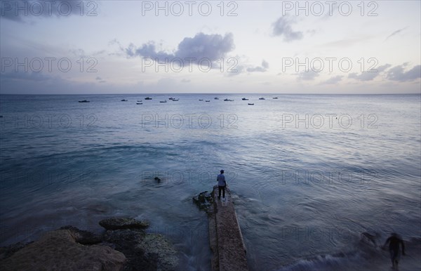 WEST INDIES, Barbados, St Lucy, Man on jetty at Half Moon Fort looking out to moored fishing boats at sunset