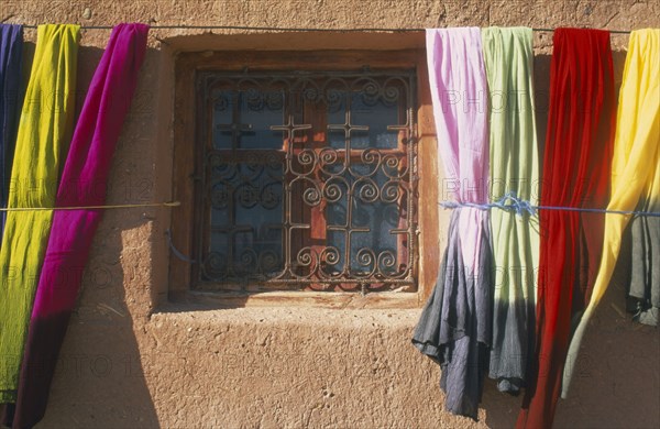 MOROCCO, Ait Benhaddou, Detail of window with decorative metal screen set into wall with colourful lengths of dyed fabric hanging at each side.