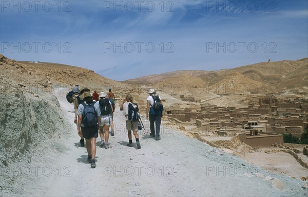 MOROCCO, High Atlas Valley, Tourist group following man on horseback along steep stony mountain path in barren landscape with rural buildings on right.